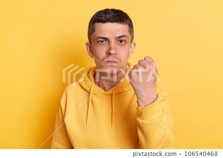 Aggressive guy. Furious quarrel. Serious strict man wearing casual hoodie showing clenched fist arguing with wife looking at camera posing isolated over yellow background. 106540468