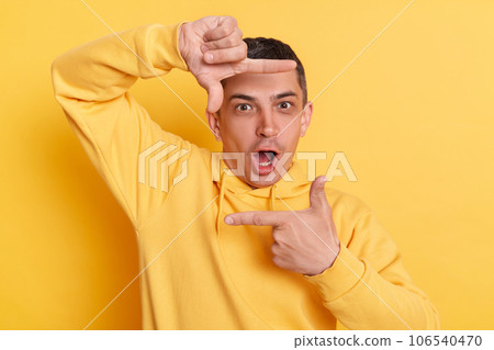 Shocked photographer. Surprised astonished man wearing casual hoodie making frame of fingers looking at camera with open mouth posing isolated over yellow background. Shocked photographer. Surprised astonished man wearing casual hoodie making frame of fingers looking at camera with open mouth posing isolated over yellow background. 106540470
