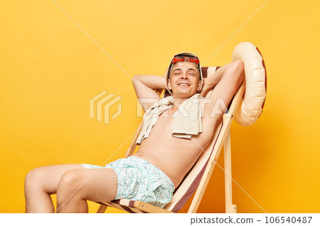 Joyful cheerful relaxed shirtless man sitting on deck chair isolated over yellow background posing with hands behind head and closed eyes relaxing at beach resting enjoying summertime. 106540487