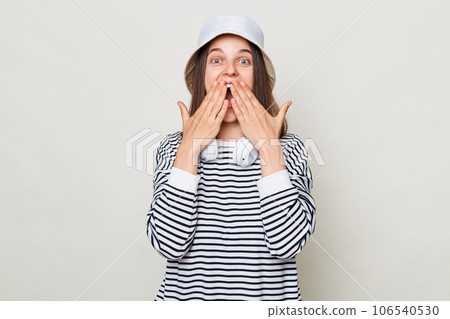 Surprised brown haired adult woman wearing striped shirt and panama standing isolated over gray background looking at camera with big eyes covering mouth with palm. 106540530