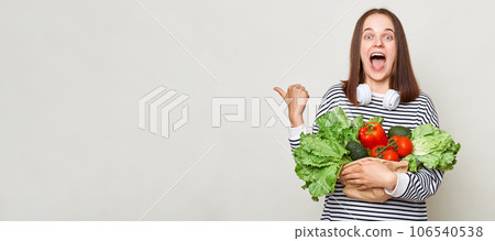 Amazed excited brown haired young woman embraces bouquet of fresh vegetables wearing striped casual shirt isolated over gray background pointing at copy space for promotion. Amazed excited brown haired young woman embraces bouquet of fresh vegetables wearing striped casual shirt isolated over gray background pointing at copy space for promotion. 106540538