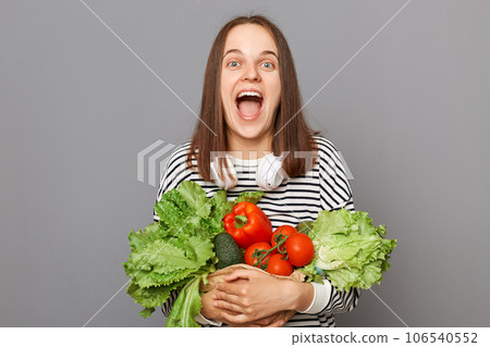 Grocery shopping for healthy eating. Shopping at the vegetarian market. Amazed excited woman embracing fresh vegetables returns from supermarket wearing striped  shirt isolated over gray background 106540552