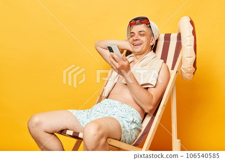 Relaxed smiling happy shirtless man sitting on deck chair isolated over yellow background using mobile phone reading news in internet checking social networks. Relaxed smiling happy shirtless man sitting on deck chair isolated over yellow background using mobile phone reading news in internet checking social networks. 106540585