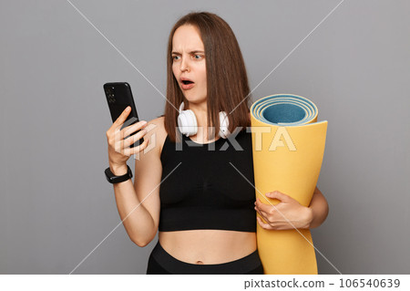 Confused shocked puzzled woman wearing black sportswear holding yoga mat standing isolated over gray background using cell phone finding yoga training in internet. Confused shocked puzzled woman wearing black sportswear holding yoga mat standing isolated over gray background using cell phone finding yoga training in internet. 106540639