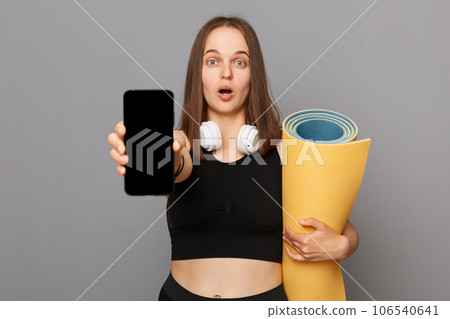 Astonished woman wearing black sportswear holding yoga mat standing isolated over gray background looking at camera with big eyes showing phone with empty screen. Astonished woman wearing black sportswear holding yoga mat standing isolated over gray background looking at camera with big eyes showing phone with empty screen. 106540641