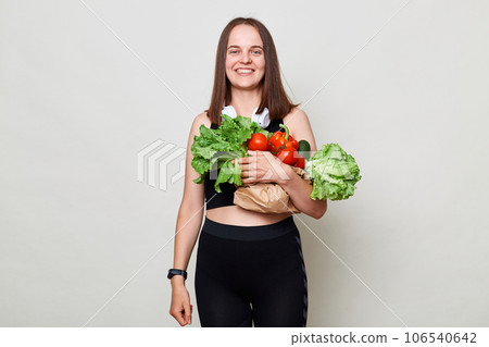 Happy smiling cheerful woman with dark hair carries vegetables bought at market isolated over gray background enjoying healthy eating looking at camera with satisfied face. Happy smiling cheerful woman with dark hair carries vegetables bought at market isolated over gray background enjoying healthy eating looking at camera with satisfied face. 106540642