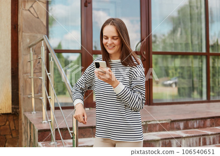 Attractive positive young beautiful caucasian woman wearing stripedshirt using smartphone standing near building reading news in internet checking social networks while walking. Attractive positive young beautiful caucasian woman wearing stripedshirt using smartphone standing near building reading news in internet checking social networks while walking. 106540651