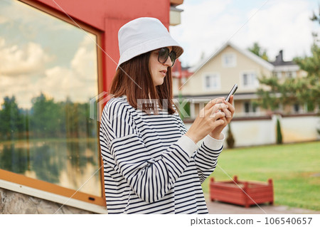 Side view of concentrated young beautiful caucasian woman wearing casual shirt and panama using smartphone standing near building window scrolling online. 106540657