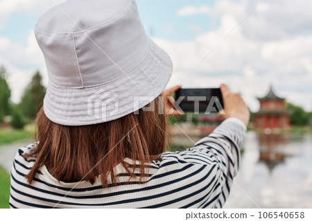 Woman in summer lake, river pond, photographing landscape on smartphone, view from back, recording a video on Internet, female in striped shirt and panama. 106540658