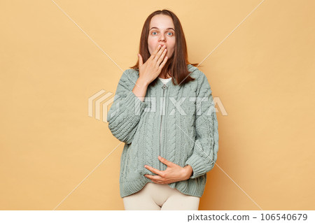 Shocked surprised young pregnant woman wearing knitted sweater standing isolated over beige background covering mouth with palm sees something scared. 106540679