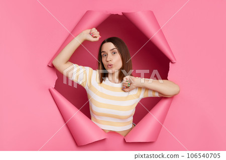 Beautiful caucasian brunette woman wearing striped t shirt posing in torn pink paper wall standing with raised arms surprised face pout lips looking directly at camera. 106540705
