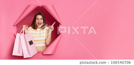 Amazed surprised caucasian young woman wearing striped shirt breaking through pink paper hole holding shopping bags pointing away at copy space for advertisement blank wall. Amazed surprised caucasian young woman wearing striped shirt breaking through pink paper hole holding shopping bags pointing away at copy space for advertisement blank wall. 106540706