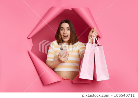 Shocked woman wearing striped shirt breaking through pink paper hole standing with shopping bags in hands using phone buying online looking at camera with open mouth and big eyes. Shocked woman wearing striped shirt breaking through pink paper hole standing with shopping bags in hands using phone buying online looking at camera with open mouth and big eyes. 106540711