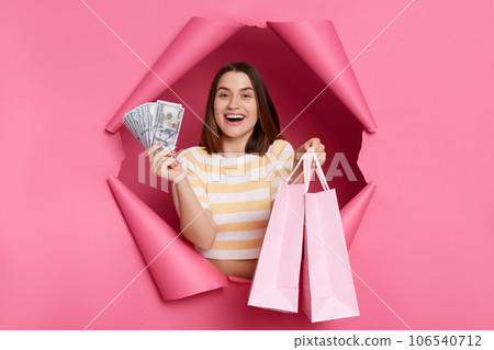 Amazed happy cheerful brunette woman wearing striped t shirt posing in torn pink paper wall showing tocamera shopping bags and dollar banknotes cash back. Amazed happy cheerful brunette woman wearing striped t shirt posing in torn pink paper wall showing tocamera shopping bags and dollar banknotes cash back. 106540712