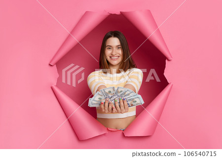 Attractive young brunette woman wearing striped t shirt posing in torn pink paper wall holding out big sum of money giving dollar banknotes rich female with cash. Attractive young brunette woman wearing striped t shirt posing in torn pink paper wall holding out big sum of money giving dollar banknotes rich female with cash. 106540715
