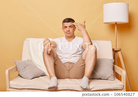 Stressed depressed brunette adult man wearing casual clothing sitting on sofa against beige wall making suicide gesture with fingers gun, looking at camera with displeased expression. 106540827