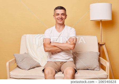 Confident positive smiling brunette young man wearing casual white t shirt sitting on sofa against beige wall posing with crossed arms looking at camera with happy expression. 106540828