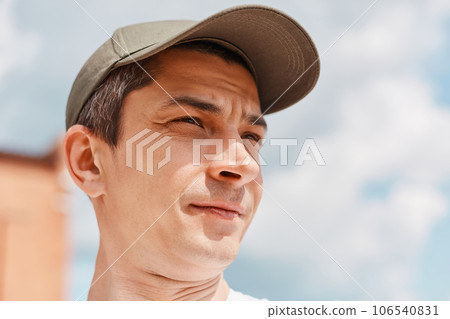 Young man posing on background of clear sky wearing baseball cap looking away with dreamy facial expression. Young man posing on background of clear sky wearing baseball cap looking away with dreamy facial expression. 106540831