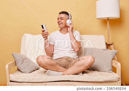 Man with smartphone listening to music on comfortable sofa against beige background enjoying favorite songs expressing positive emotions smiling happily. Man with smartphone listening to music on comfortable sofa against beige background enjoying favorite songs expressing positive emotions smiling happily. 106540834