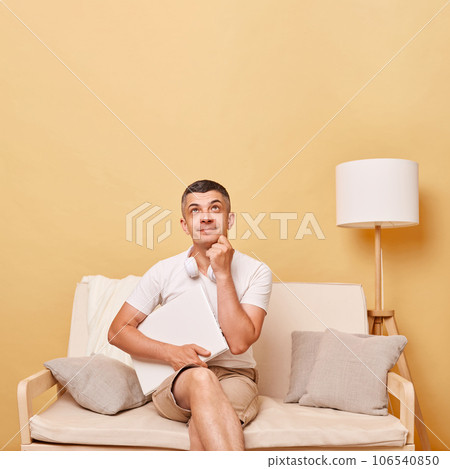 Pensive thoughtful adult man wearing white t-shirt and shorts sitting on sofa with closed laptop against beige wall looking upwards at advertisement area copy space for promotion. Pensive thoughtful adult man wearing white t-shirt and shorts sitting on sofa with closed laptop against beige wall looking upwards at advertisement area copy space for promotion. 106540850