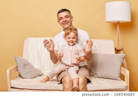Attractive man with his daughter clapping hands and laughing while sitting on sofa tohgether playing games having fun at home expressing happiness. Attractive man with his daughter clapping hands and laughing while sitting on sofa tohgether playing games having fun at home expressing happiness. 106540885