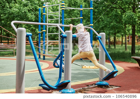 Full length portrait of little girl wearing baseball cap doing physical exercises on sportsground dressed casual closing making training on legs on on outdoor simulator. 106540896