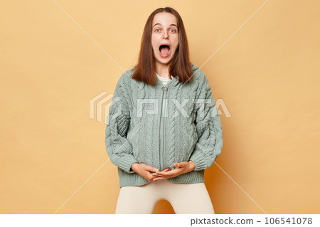 Shocked young pregnant woman wearing knitted sweater standing isolated over beige background holding her belly starting contractions screaming with widely open mouth. 106541078