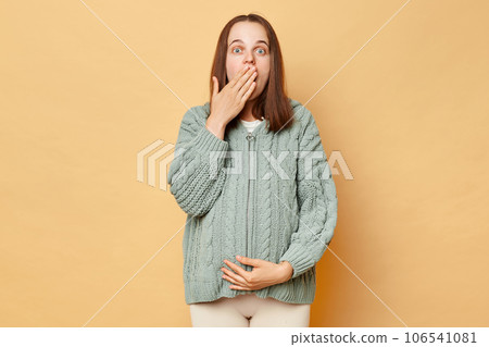 Shocked amazed pregnant woman wearing warm knitted sweater touching belly posing over beige background covering mouth with palm sees something astonished. Shocked amazed pregnant woman wearing warm knitted sweater touching belly posing over beige background covering mouth with palm sees something astonished. 106541081