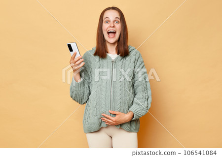 Happy excited positive pregnant woman wearing knitted sweater standing isolated beige background holding cell phone looking at camera screaming with happiness. Happy excited positive pregnant woman wearing knitted sweater standing isolated beige background holding cell phone looking at camera screaming with happiness. 106541084