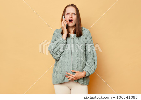 Shocked astonished pregnant woman wearing warm knitted sweater touching belly posing over beige background talking on mobile phone looking at camera with surprised face. Shocked astonished pregnant woman wearing warm knitted sweater touching belly posing over beige background talking on mobile phone looking at camera with surprised face. 106541085