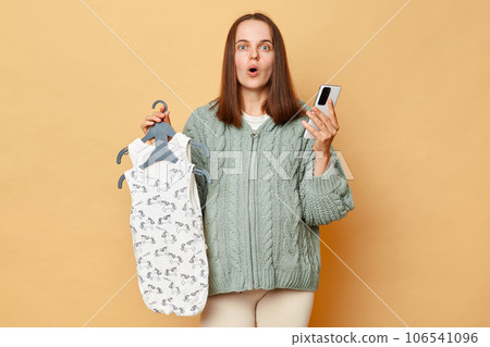 Shocked amazed young pregnant woman with baby bodysuits isolated over beige background using mobile phone ordering clothing for her future child looking at camera with big eyes. 106541096