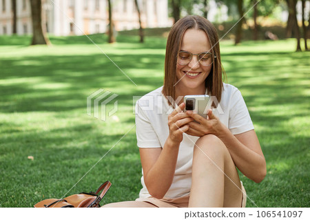 Smiling cheerful positive woman wearing casual white t-shirt sitting in park on green grass using mobile phone chatting with friends checking social networks. 106541097