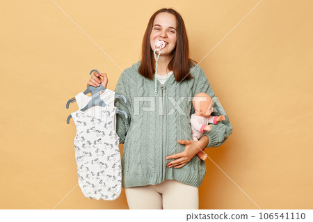 Positive funny pregnant woman with nipple in mouth wearing knitted sweater holding baby's bodysuits and doll standing isolated beige background looking at camera with happy face. Positive funny pregnant woman with nipple in mouth wearing knitted sweater holding baby's bodysuits and doll standing isolated beige background looking at camera with happy face. 106541110