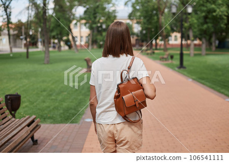Back view of brown haired girl resting strolling pastime on fresh air outdoors wearing white t-shirt and holding leather backpack walking on path in city park. Back view of brown haired girl resting strolling pastime on fresh air outdoors wearing white t-shirt and holding leather backpack walking on path in city park. 106541111