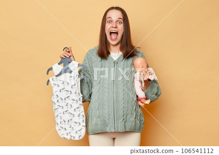 Overjoyed excited amazed young pregnant woman with baby bodysuits and doll isolated over beige background looking at camera and screaming with happy satisfied face. Overjoyed excited amazed young pregnant woman with baby bodysuits and doll isolated over beige background looking at camera and screaming with happy satisfied face. 106541112