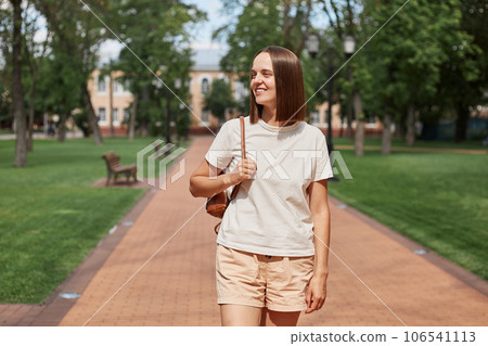 Attractive brown haired girl resting strolling pastime on fresh air outdoors wearing white t-shirt and holding backpack walking on path in city park. Attractive brown haired girl resting strolling pastime on fresh air outdoors wearing white t-shirt and holding backpack walking on path in city park. 106541113