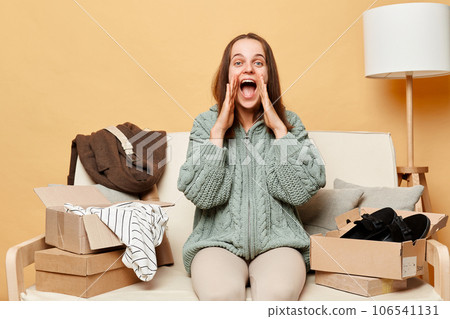 Excited caucasian woman sitting on sofa among boxes with clothing against beige wall screaming with widely open mouth making announcement get her order from store. Excited caucasian woman sitting on sofa among boxes with clothing against beige wall screaming with widely open mouth making announcement get her order from store. 106541131