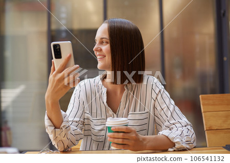 Happy university student girl using mobile phone and drinking coffee smiling while looking away, businesswoman having break enjoying hot beverage in cafe. 106541132