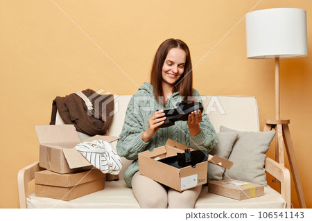Attractive happy positive woman sitting on sofa among boxes with clothing against beige wall buying new shoes admiring unpacking package from shop delivery. Attractive happy positive woman sitting on sofa among boxes with clothing against beige wall buying new shoes admiring unpacking package from shop delivery. 106541143
