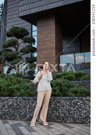 Full length portrait of attractive woman in elegant blouse and trousers standing in street and talking on mobile phone looking smiling away having business conversation. 106541154