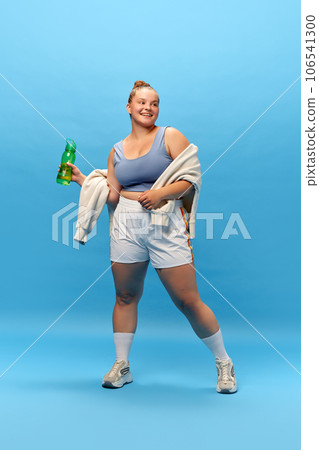Full length portrait of young smiling plus-size woman staying in sportswear with bottle for water against blue studio background. rest. energy. 106541300