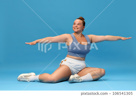 Self-care and well being. Young overweight woman stretching on floor in sportswear against blue studio background. Motivation. 106541305