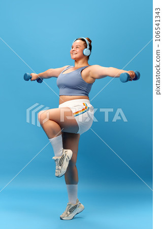 Full length photo of smiling overweight woman wearing sportswear training, exercising with dumbbells in headphones with music. standing leg raises. 106541343