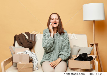Attractive laughing woman sitting on sofa among boxes with clothing against beige wall talking on smartphone with friend saying about internet shopping. Attractive laughing woman sitting on sofa among boxes with clothing against beige wall talking on smartphone with friend saying about internet shopping. 106541349
