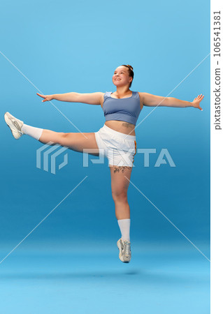 Full length photo of smiling overweight woman wearing sportswear jumping and doing exercises isolated on studio blue background. 106541381