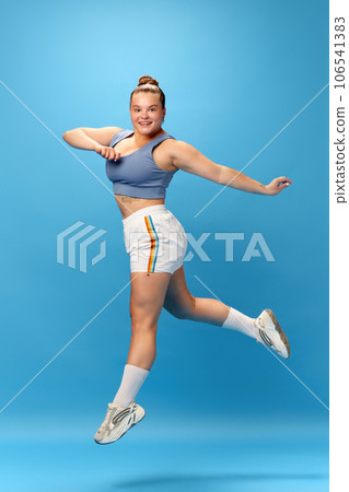 Full length portrait of young smiling fat overweight woman wearing sportswear doing exercise, jumping isolated on studio blue background. 106541383
