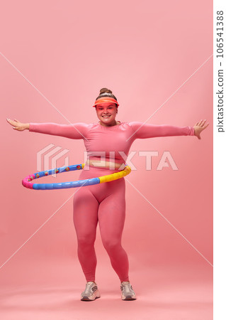 Full lenght portrait of young overweight woman dressed sporty training, warm up with hula hoop isolated on pink studio background. 106541388