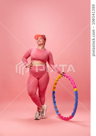 Full lenght portrait of young overweight woman dressed sporty doing exercises with hula hoop isolated on pink studio background. 106541389