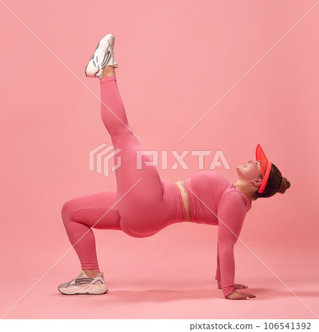 Full body side view of young overweight sporty woman in pink top and legging, training legs and back isolated studio background. Single leg bridge. Full body side view of young overweight sporty woman in pink top and legging, training legs and back isolated studio background. Single leg bridge. 106541392