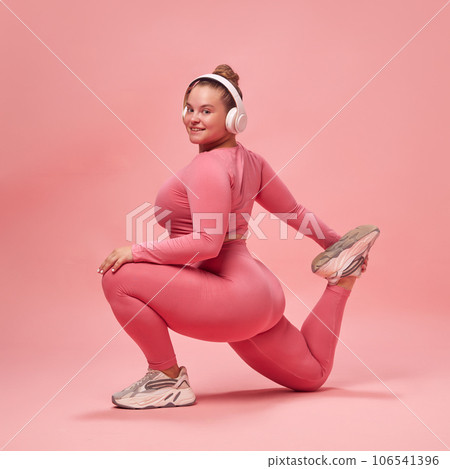 Full body portrait of young chubby sporty woman in pink top and legging, training legs with music in headphones isolated studio background. 106541396